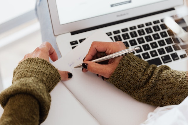 A person writing something on a notebook before a laptop